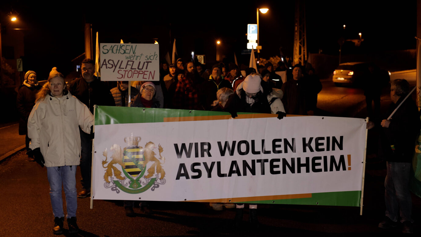 Anhänger der Freien Sachsen laufen über eine dunkle Straße und halten ein Banner mit der Aufschrift "Wir wollen kein Asylbewerberheim" hoch – ein Symbolbild für die geemeinsame Abstimmung zwischen CDU, Linkspartei und Freien Sachsen