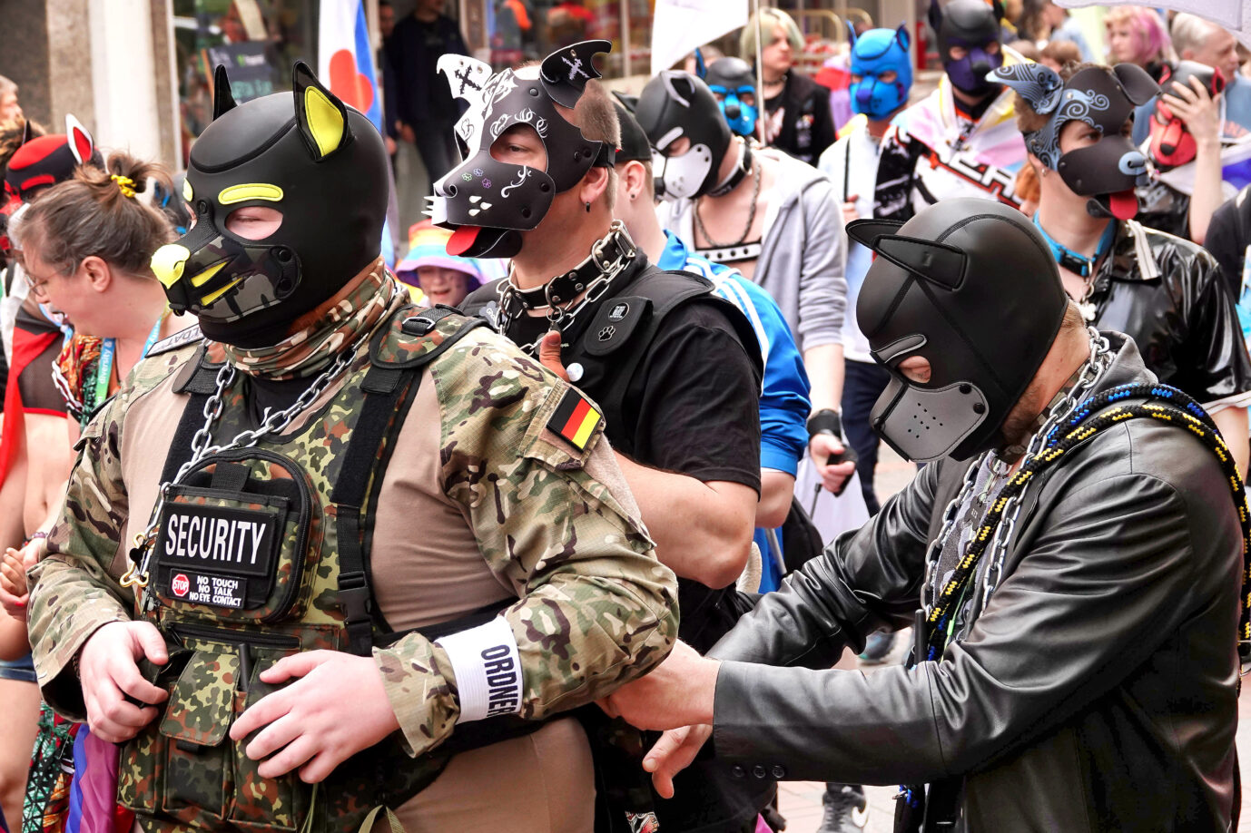 Eine Gruppe von Menschen nimmt an einer queeren Demonstration oder einem CSD (Christopher Street Day) teil. Im Vordergrund stehen mehrere Personen in sogenannten „Pup-Play“-Outfits: Sie tragen schwarze, hundeähnliche Masken mit aufgesetzten Ohren und zum Teil farbigen Details. Viele sind mit Halsbändern, Ketten, Leder- oder Neoprenkleidung sowie Accessoires aus der BDSM-Szene ausgestattet. Eine Person vorne links trägt eine Tarnuniform mit einer schwarz-rot-goldenen Deutschlandflagge auf dem Ärmel, einer Weste mit dem Aufdruck „Security“ und einer Armbinde mit der Aufschrift „Ordner“. Auf ihrer Brust ist außerdem ein Schild befestigt mit den Hinweisen „No touch – No talk – No eye contact“. Um sie herum befinden sich weitere Personen in bunter oder freizügiger Kleidung. Im Hintergrund ist eine größere Menschenmenge sichtbar. Die Szene spielt sich in einer belebten Innenstadt ab und vermittelt eine Mischung aus Demonstration, Performance und öffentlichem queeren Ausdruck. Trotz tierisch guter Sicherheitskräfte mußte der CSD-Umzug abgesagt werden: Polizei schweigt über konkrete Gründe. Foto: IMAGO / Funke Foto Services