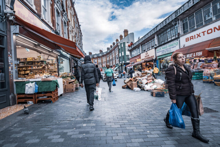 Ein Mann und eine Frau gehen auf einer grauen trostlosen Straße aneinander vorbei – es handelt sich um eine Szene aus dem Londoner Stadtteil Brixton – über 50 Prozent der Bewohner haben hier einen Migrationshintergrund – Symbolbild dafür, daß sich viele Briten fremd im Land fühlen