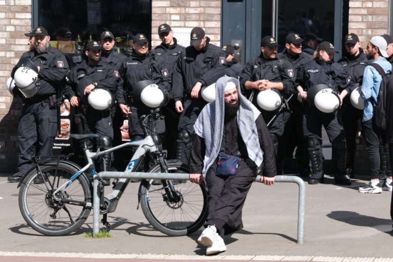 Ein Mann mit traditionell islamischer Kleidung sitzt vor einer Gruppe von Polizisten in Einsatzkleidung und Helmen. Im Vordergrund steht ein Fahrrad, im Hintergrund lehnen sich die Polizisten an eine Hauswand und beobachten das Geschehen. Islamisten im Visier: 2024 liefen deutlich mehr Verfahren gegen sie als gegen Links- und Rechtsextreme. Foto: IMAGO / epd