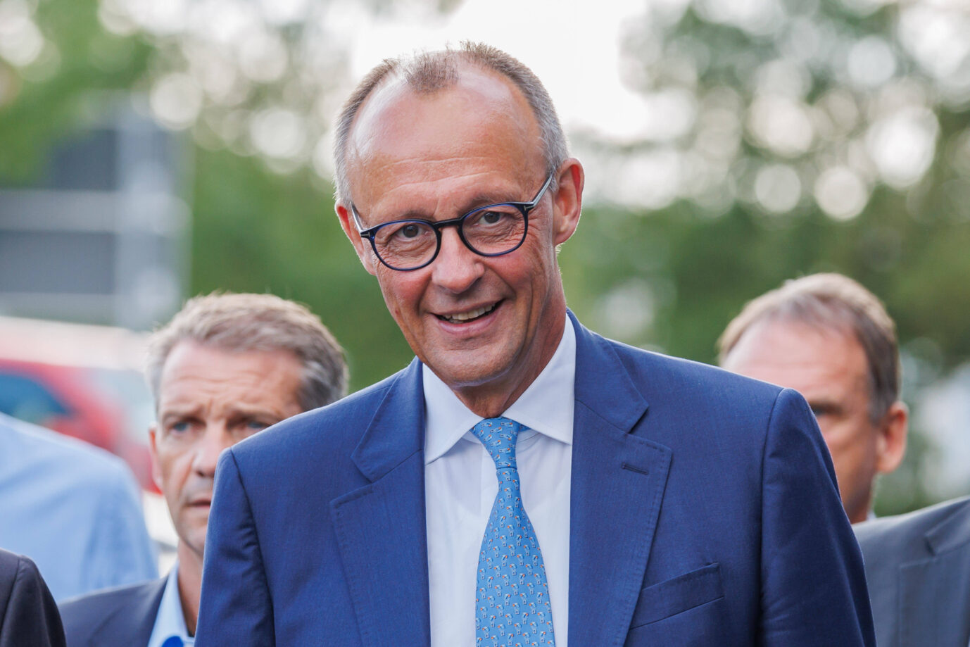 Friedrich Merz, lächelnd im blauen Anzug und heller Krawatte, steht im Freien. Er trägt eine runde Brille. Im Hintergrund sind weitere Männer sowie verschwommene Bäume und Autos zu erkennen