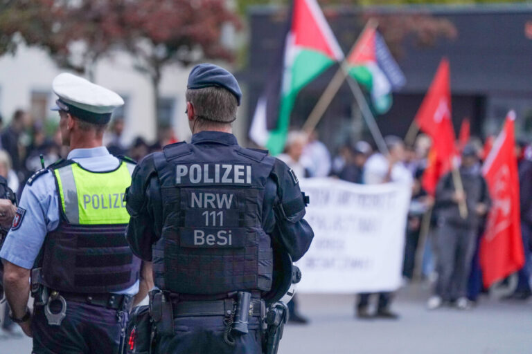 Zwei Polizisten – einer in Uniform mit neongelber Weste, einer in Einsatzkleidung mit „Polizei NRW BeSi“-Aufschrift – stehen mit dem Rücken zur Kamera vor einer Gruppe von Demonstrierenden. Im Hintergrund sind palästinensische und türkische Fahnen sowie ein weißes Transparent mit arabischer oder englischer Aufschrift zu sehen. Polizisten sichern eine Demonstration mit islamistischem Hintergrund – viele Gefährder leben laut Bundesregierung weiterhin in Deutschland. Foto: IMAGO / Steinsiek.ch