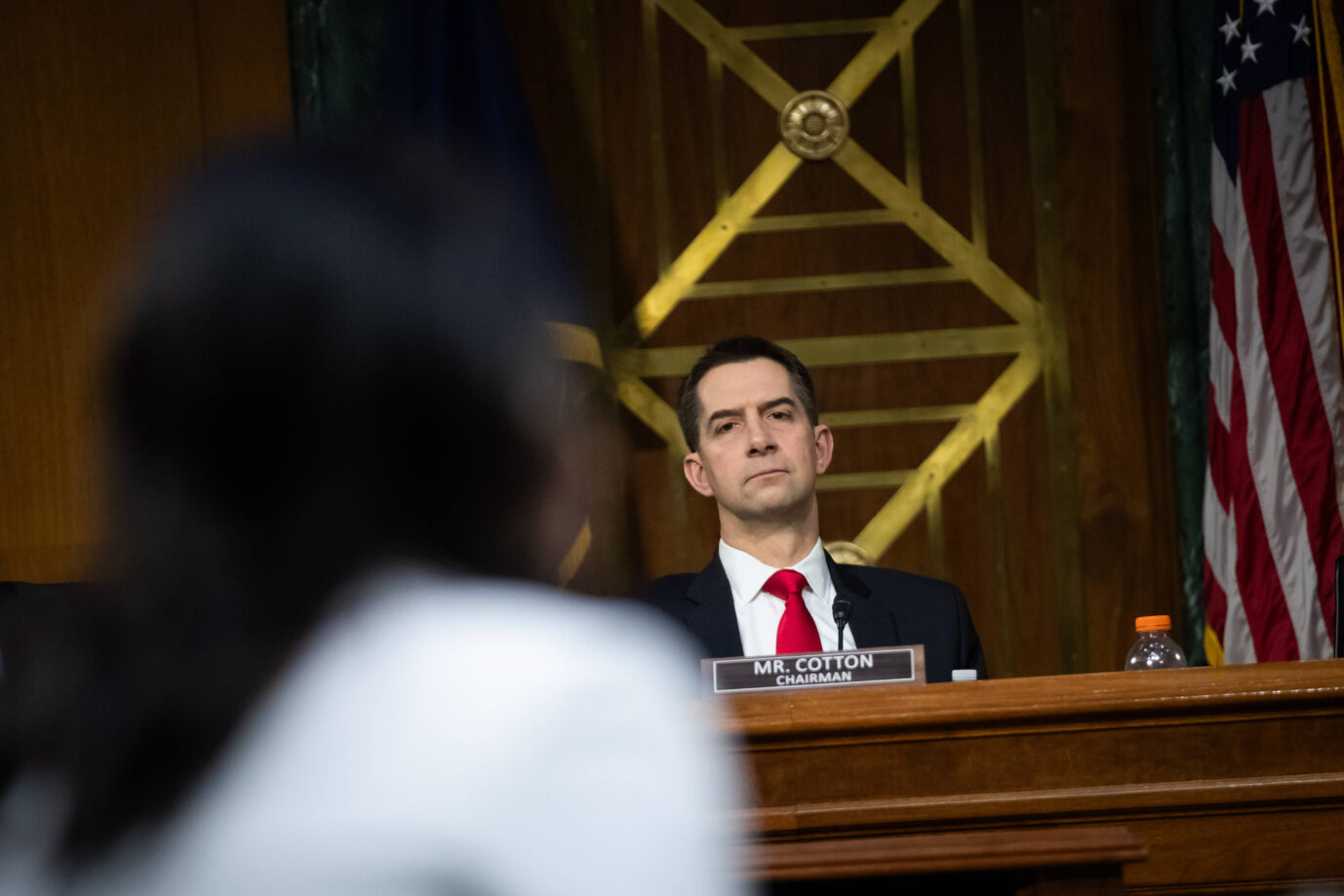 S-Senator Tom Cotton (Republikaner, Arkansas) sitzt mit ernster Miene als Vorsitzender des Geheimdienstausschusses im Senatssaal, vor ihm unscharf im Vordergrund eine weibliche Person; im Hintergrund sind das Wappen des Senats und eine US-Flagge zu sehen.