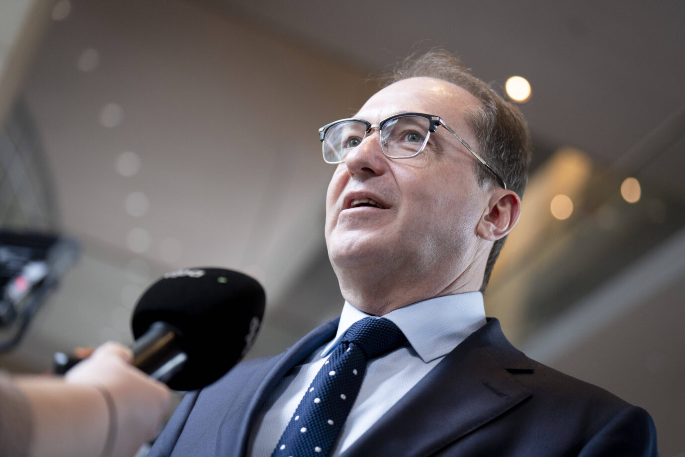Ein Mann im Anzug spricht in ein Mikrofon. Er trägt eine Brille mit transparentem Rahmen, ein weißes Hemd und eine dunkelblaue Krawatte mit weißen Punkten. Die Kamera blickt leicht von unten auf ihn, im Vordergrund ist ein schwarzes Mikrofon mit Senderlogo sichtbar. Im Hintergrund sind unscharf Lichtpunkte und Architekturdetails eines modernen Gebäudes zu erkennen. Deutschlands Innenminister Alexander Dobrindt (CSU) ordnet an seinem zweiten Tag Zurückweisungen an. Foto: IMAGO / Bernd Elmenthaler