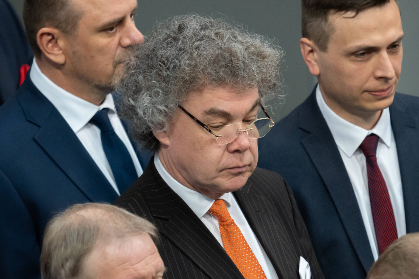 Das Bild zeigt eine Gruppe von Männern in Anzügen, vermutlich bei einer offiziellen Veranstaltung oder einem parlamentarischen Anlass. Im Zentrum steht ein Mann mit auffällig grauem, krausem Haar und randloser Brille, die tief auf der Nase sitzt. Er trägt einen dunklen Nadelstreifenanzug, ein weißes Hemd und eine orangefarbene Krawatte mit kleinen weißen Punkten. Er hat die Augen geschlossen oder niedergeschlagen und wirkt konzentriert oder in sich gekehrt. Um ihn herum stehen mehrere Männer in dunklen Anzügen, weißen Hemden und Krawatten, einer mit blauer, ein anderer mit roter Krawatte. Die Personen stehen eng beieinander, möglicherweise bei einer Gedenkminute oder in Vorbereitung auf ein Gruppenfoto. Der Hintergrund ist neutral und lässt auf ein institutionelles Umfeld wie ein Parlament oder eine Regierungsveranstaltung schließen. Matthias Moosdorf, umgeben von seinen Fraktionskollegen im Bundestag: Er vertritt die AfD nicht mehr im Außenausschuß. Foto: IMAGO / Bihlmayerfotografie