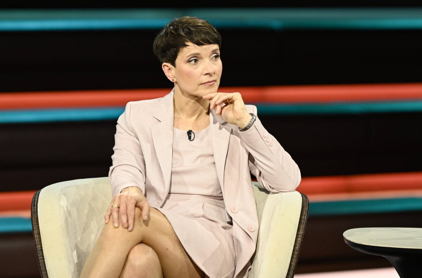 Auf dem Bild ist Frauke Petry in einem Fernsehstudio zu sehen. Sie sitzt in einem hell gepolsterten Sessel mit dunklen Armlehnen. Petry trägt ein blassrosafarbenes Kostüm, bestehend aus einem Blazer und einem knielangen Kleid. Ihre Beine sind übereinandergeschlagen, die linke Hand liegt locker auf dem rechten Bein. Mit der rechten Hand stützt sie ihr Kinn, der Blick ist konzentriert und leicht nach rechts gewandt, als würde sie jemandem zuhören oder über etwas nachdenken. Im Hintergrund des Studios verlaufen waagerechte, farbige Streifen in Schwarz, Rot und Türkisblau. Der Gesamteindruck ist ruhig und sachlich, Petrys Körperhaltung wirkt aufmerksam und nachdenklich. Auf ihrem Blazer ist ein Mikrofon angebracht, was darauf hinweist, daß sie Teil einer TV-Diskussion oder Talkrunde ist.