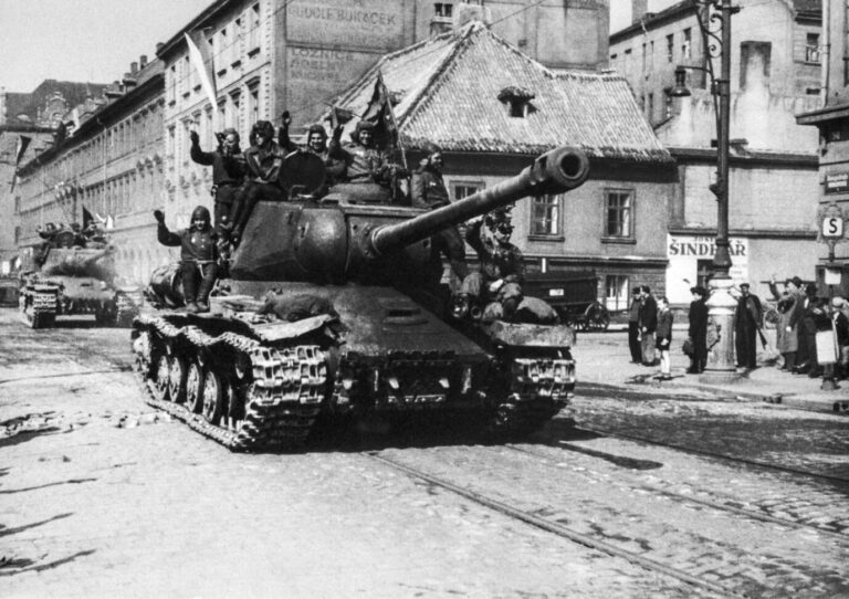 Schwarz-weiß-Fotografie aus dem Mai 1945: Ein sowjetischer Panzer vom Typ IS-2 fährt durch eine Straße in Prag. Auf dem Panzer sitzen und stehen mehrere rotarmistische Soldaten, die lachend in die Kamera winken oder mit erhobener Faust jubeln. Zivilisten am Straßenrand begrüßen sie, einige heben ebenfalls die Arme zum Gruß. Im Hintergrund sind historische Gebäude mit beschädigten Dächern und Fassaden zu sehen, teils mit tschechischer Werbung bemalt. Die Szene vermittelt den Moment des Einmarschs der Roten Armee in die tschechische Hauptstadt kurz vor Kriegsende.