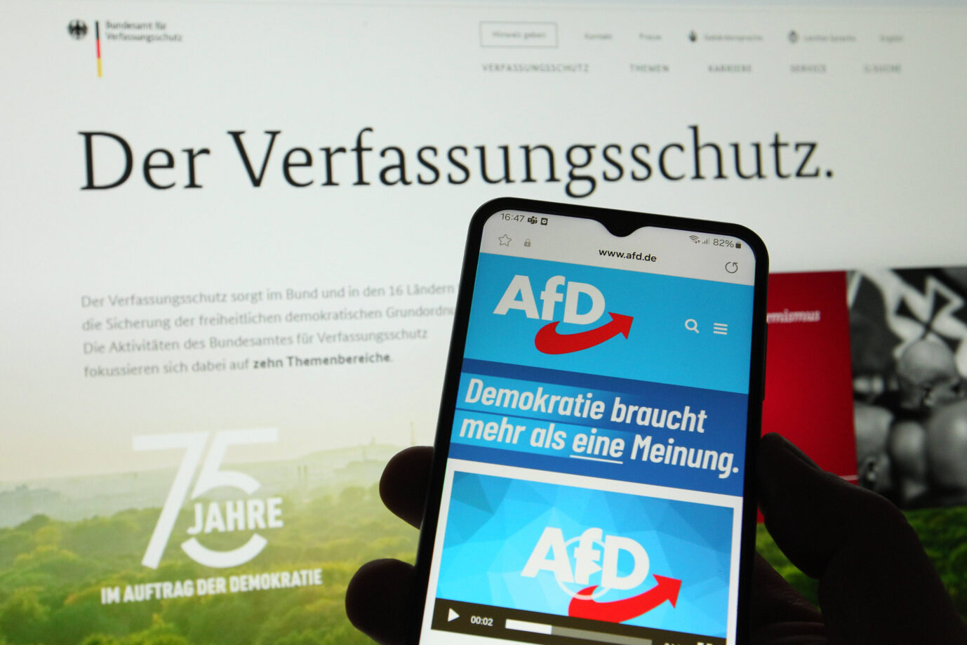 Eine Hand hält ein Handy, auf dem die Person offenbar die Webseite der AfD aufgerufen hat. Im Hintergrund ist die Webseite des Verfassungsschutzes zu sehen – tritt die Person etwa in die AfD ein?