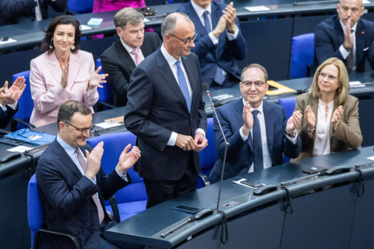 Friedrich Merz steht im Plenarsaal des Bundestages an seinem Platz, umgeben von applaudierenden Abgeordneten der CDU/CSU-Fraktion, darunter Jens Spahn, Dorothee Bär und Alexander Dobrindt.