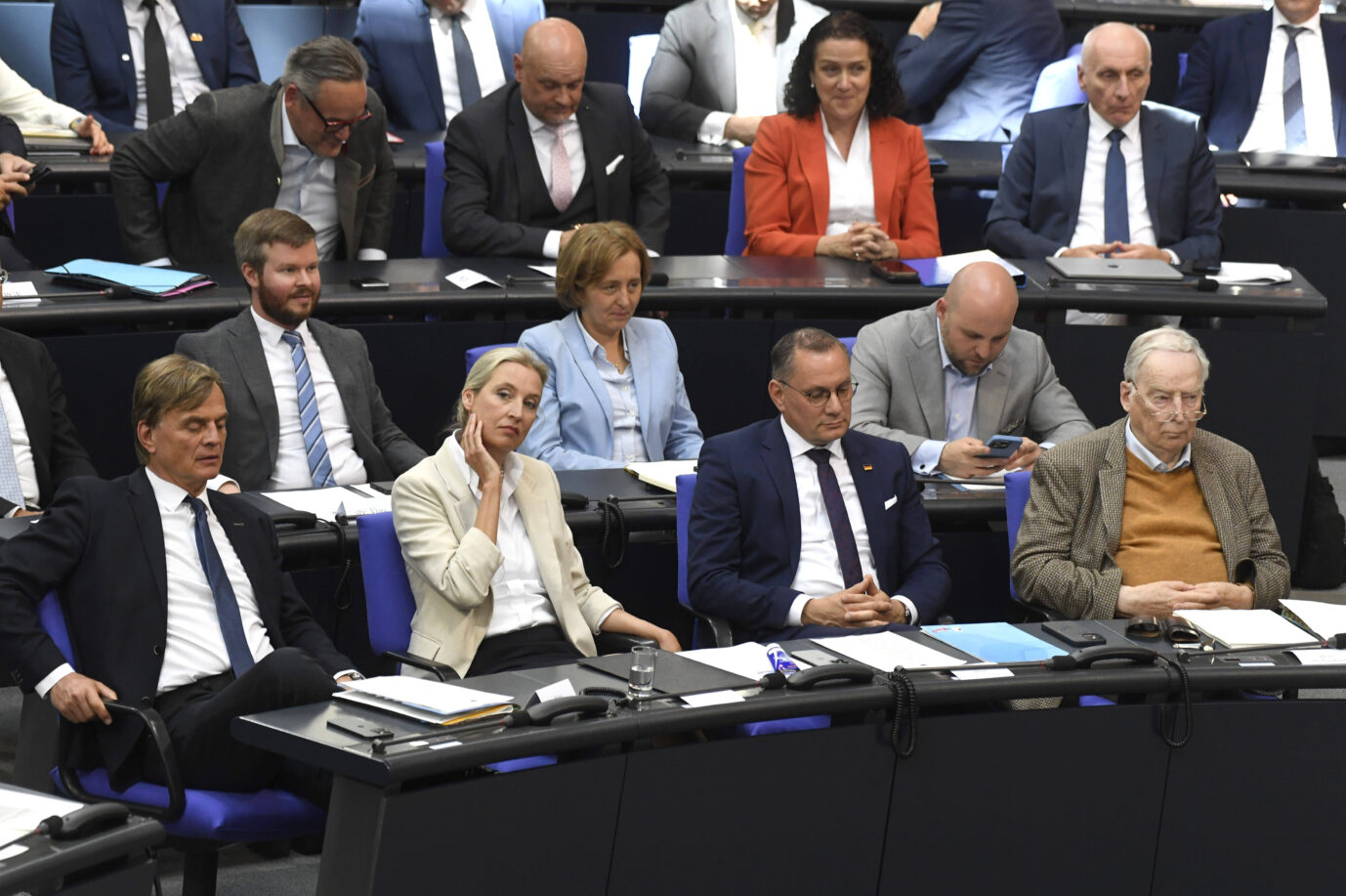 Mehrere Abgeordnete der AfD-Fraktion sitzen im Plenarsaal des Deutschen Bundestags. In der ersten Reihe befinden sich unter anderem Alice Weidel mit nachdenklicher Miene, Tino Chrupalla mit gefalteten Händen und Alexander Gauland in braunem Sakko und senfgelbem Pullover. Neben ihnen sitzen weitere Fraktionsmitglieder, viele wirken konzentriert oder ernst, einer blickt auf sein Smartphone. Die Sitzung läuft, auf den Tischen liegen Unterlagen, Mikrofone und Aktenmappen.