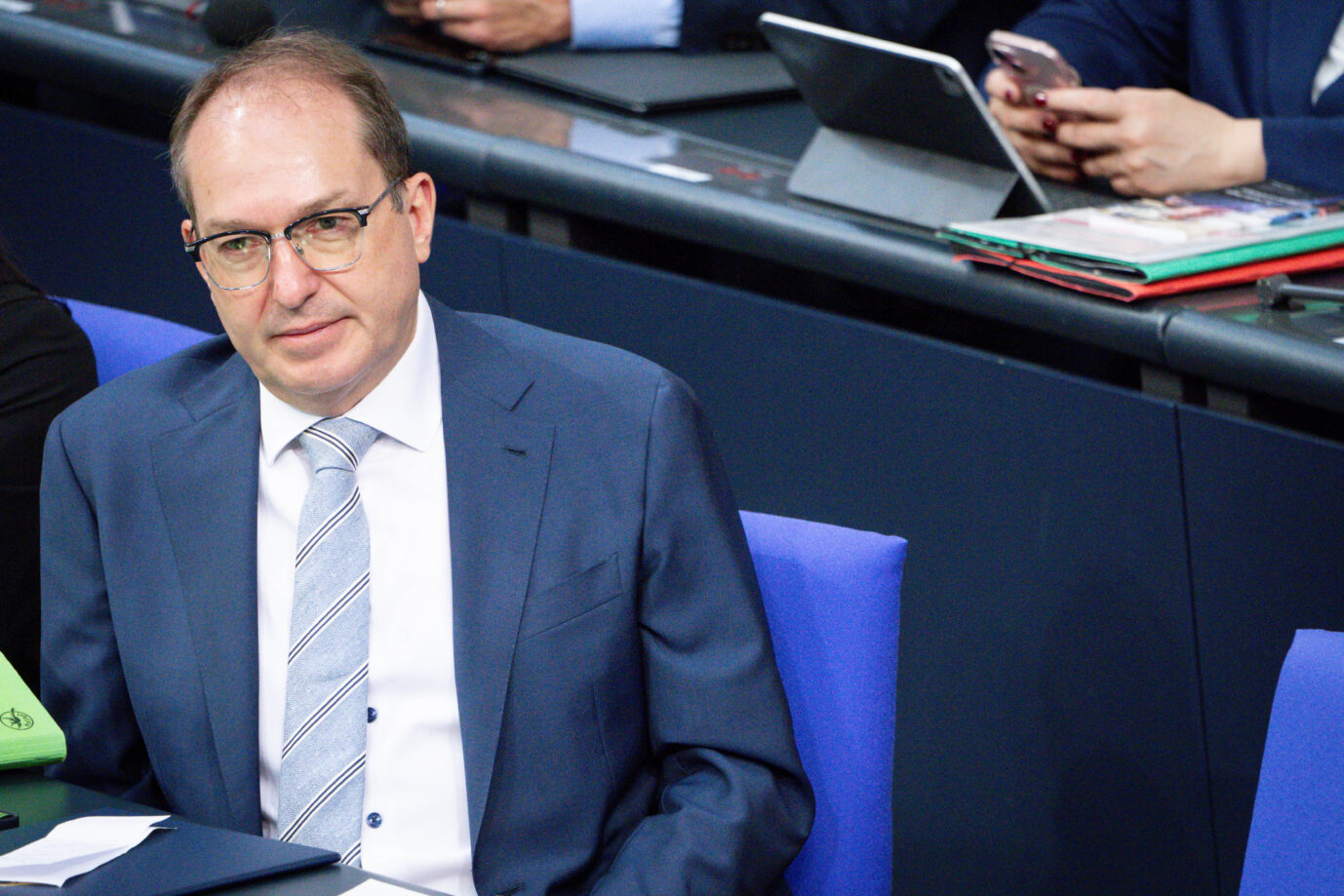 Alexander Dobrindt (CSU) sitzt im Plenarsaal des Bundestages, trägt einen blauen Anzug mit gestreifter Krawatte und blickt nachdenklich zur Seite. Im Hintergrund sind weitere Abgeordnete mit Tablets und Unterlagen zu sehen.