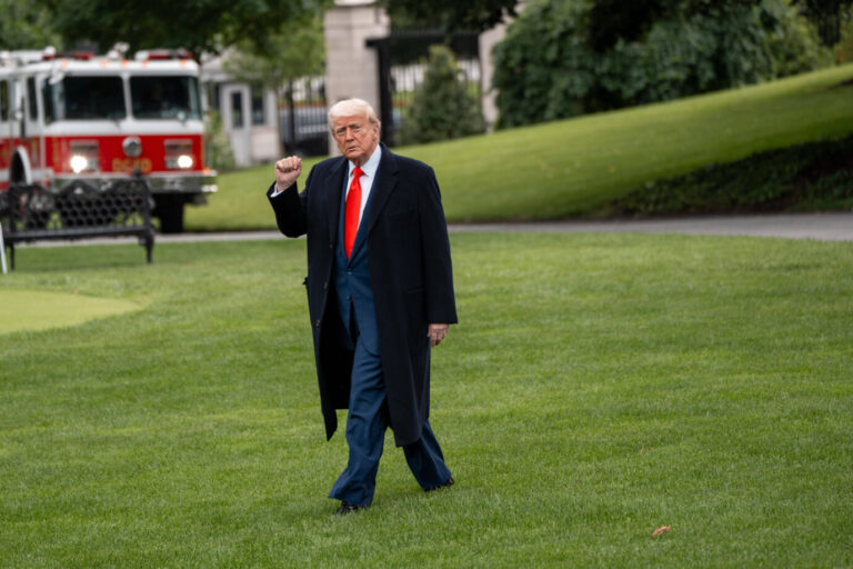 Ein älterer Mann mit blondem Haar, bekleidet mit einem dunklen Mantel, blauem Anzug und roter Krawatte, geht über eine Rasenfläche und hebt die rechte Faust leicht in die Luft. Im Hintergrund steht ein rotes Feuerwehrauto vor einem Gebäude mit Bäumen und Rasenflächen. US-Präsident Donald Trump will Stärke zeigen: Gegen die EU sollen Strafzölle in Höhe von 50 Prozent gelten. Foto: IMAGO / ZUMA Press Wire