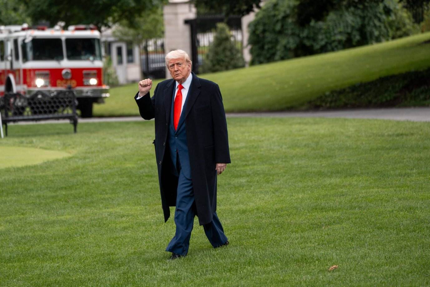 Ein älterer Mann mit blondem Haar, bekleidet mit einem dunklen Mantel, blauem Anzug und roter Krawatte, geht über eine Rasenfläche und hebt die rechte Faust leicht in die Luft. Im Hintergrund steht ein rotes Feuerwehrauto vor einem Gebäude mit Bäumen und Rasenflächen. US-Präsident Donald Trump will Stärke zeigen: Gegen die EU sollen Strafzölle in Höhe von 50 Prozent gelten. Foto: IMAGO / ZUMA Press Wire