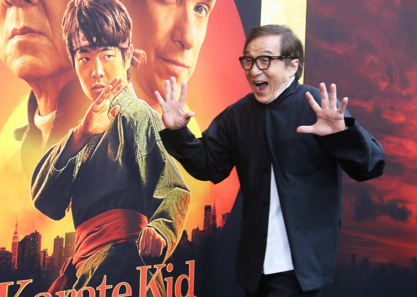 Schauspieler Jackie Chan posiert lachend und mit ausgestreckten Händen vor einem Filmplakat von „Karate Kid – Legends“, das ihn sowie weitere Darsteller in Kampfhaltung zeigt. Jackie Chan macht Spaß bei der Premiere von „Karate Kid – Legends“, in dem er erneut als Kung-Fu-Mentor zu sehen ist. Foto: IMAGO / Cover-Images