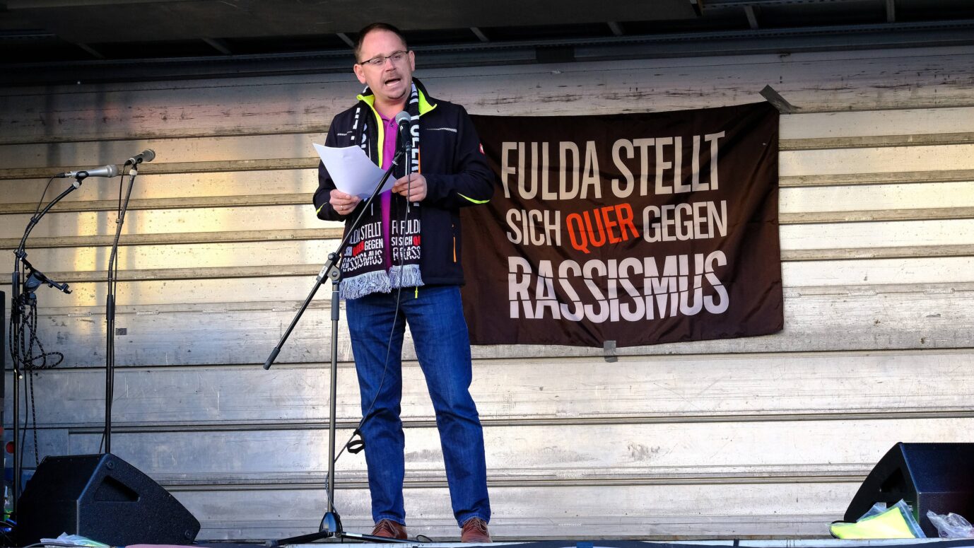 Ein Mann mit einem Schal steht auf einer Bühne – hinter ihm weht die Fahne des linksradikalen Bündnisses "Fulda stellt sich quer" – die AfD wird nun unter Umständen deren Wonhraum übernehmen
