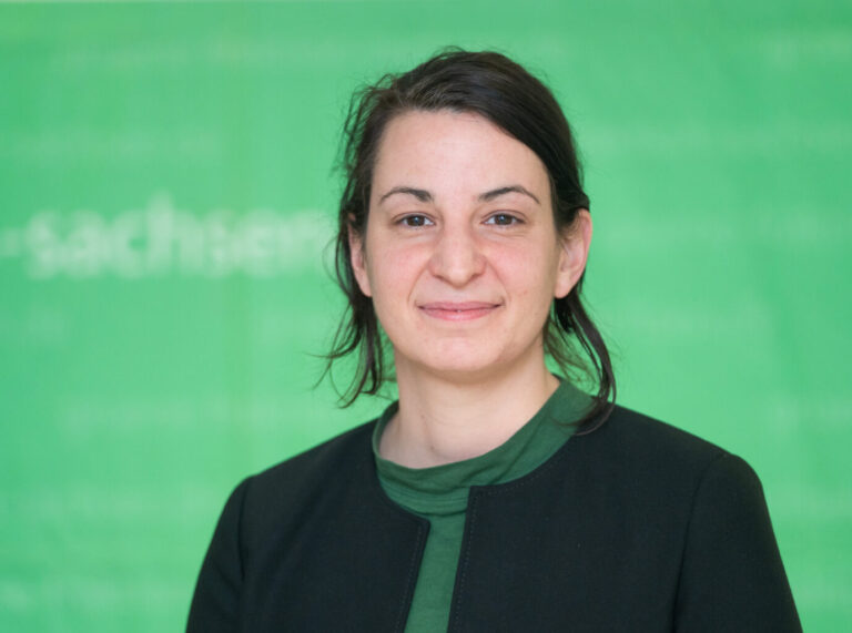 Porträtfoto der sächsischen Landtagsabgeordneten der Grünen, Christin Melcher, aufgenommen vor einem unscharf grünen Hintergrund mit der teils lesbaren Aufschrift „sachsen“; die Politikerin trägt ein dunkles Jackett über einem grünen Oberteil und blickt mit leichtem Lächeln direkt in die Kamera; das Bild eignet sich zur Illustration politischer Berichterstattung über Bündnis 90/Die Grünen in Sachsen, insbesondere im Kontext von linksextremen Angriffen auf Abgeordnetenbüros, innerparteilichen Debatten oder sicherheitspolitischen Themen. Grünen-Abgeordnete Melcher: Zwischen linker Gewalt und linkem Selbstverständnis. Foto: picture alliance/dpa | Ronald Bonß