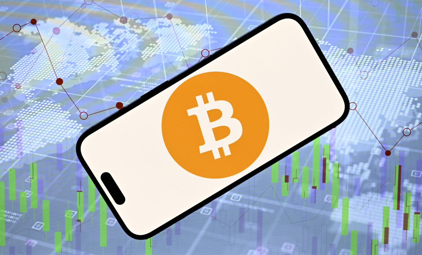 Das Bild zeigt das „Bitcoin“-Logo vor einer digitalen Weltkarte. Es ist ein Symbolbild für Cryptowährungen und das globale Geldsystem.