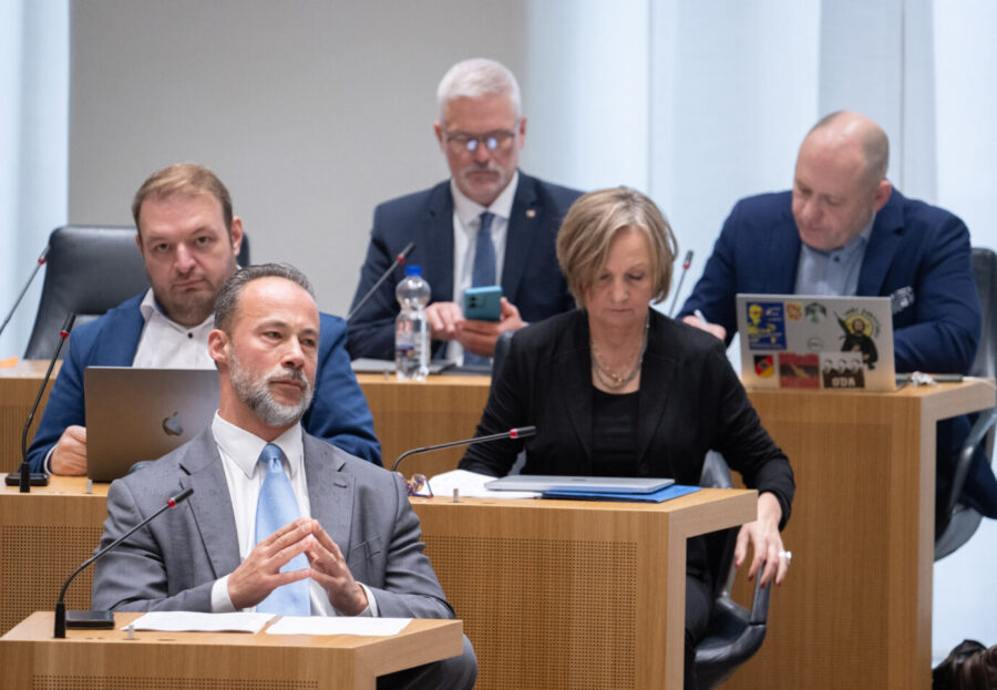 Fünf Abgeordnete sitzen im Plenarsaal des Landtags Rheinland-Pfalz. Im Vordergrund sitzt ein Mann im grauen Anzug mit hellblauer Krawatte, die Hände gefaltet. Hinter ihm arbeiten zwei Männer an Laptops, eine Frau schaut auf Unterlagen, ein weiterer Mann tippt auf sein Smartphone. Alle tragen formelle Kleidung. Jan Bollinger (l,AfD), Fraktionsvorsitzender seiner Partei, verfolgt eine Debatte im Landtag von Rheinland-Pfalz. Bald ohne Mitarbeiter? Die AfD-Fraktion im Mainzer Landtag. Foto: picture alliance/dpa | Boris Roessler