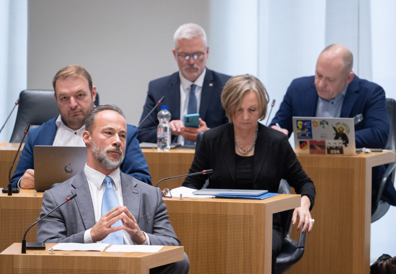 Fünf Abgeordnete sitzen im Plenarsaal des Landtags Rheinland-Pfalz. Im Vordergrund sitzt ein Mann im grauen Anzug mit hellblauer Krawatte, die Hände gefaltet. Hinter ihm arbeiten zwei Männer an Laptops, eine Frau schaut auf Unterlagen, ein weiterer Mann tippt auf sein Smartphone. Alle tragen formelle Kleidung. Jan Bollinger (l,AfD), Fraktionsvorsitzender seiner Partei, verfolgt eine Debatte im Landtag von Rheinland-Pfalz. Bald ohne Mitarbeiter? Die AfD-Fraktion im Mainzer Landtag. Foto: picture alliance/dpa | Boris Roessler