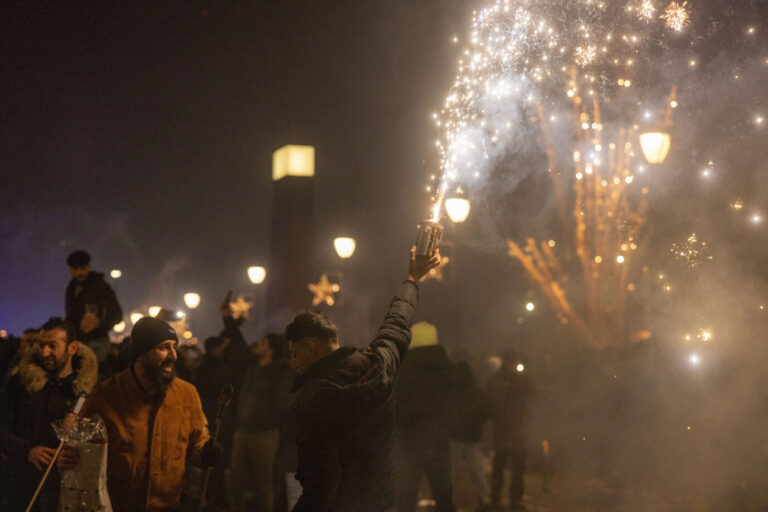 Eine größere Menschengruppe feiert nachts im Freien mit Feuerwerk. Im Vordergrund hält ein junger Mann eine zischende Feuerwerksbatterie senkrecht in die Luft, Funken sprühen. Andere Personen lachen, unterhalten sich oder tragen weitere Feuerwerkskörper. Die Szene ist von Laternenlicht und Rauch durchzogen, im Hintergrund sind Lichter und Silhouetten zu erkennen. Zehntausende Asylbewerber zelebrieren am 1. Januar ihren Geburtstag. Die Bundesregierung nennt dafür praktische Gründe – der AfD-Abgeordnete Curio äußert Skepsis.