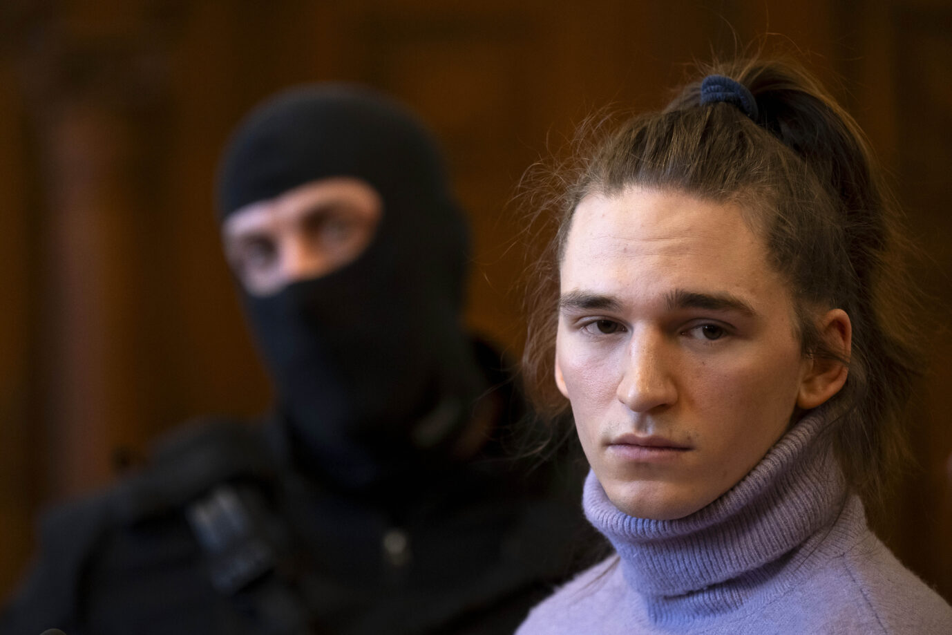 Ein junger Mann mit langen Haaren, die zu einem Zopf gebunden sind, sitzt in einem Gerichtssaal und blickt ernst. Er trägt einen lilafarbenen Rollkragenpullover. Im unscharfen Hintergrund steht eine vermummte Person in schwarzer Kleidung, vermutlich ein Beamter mit Gesichtshaube, der die Szene bewacht. Die Stimmung wirkt angespannt und konzentriert. Linksextremist Simeon „Maja“ T. mit Rollkragen vor einem ungarischen Gericht: Damit niemand den Adamsapfel sieht? Foto: picture alliance / ASSOCIATED PRESS | Denes Erdos