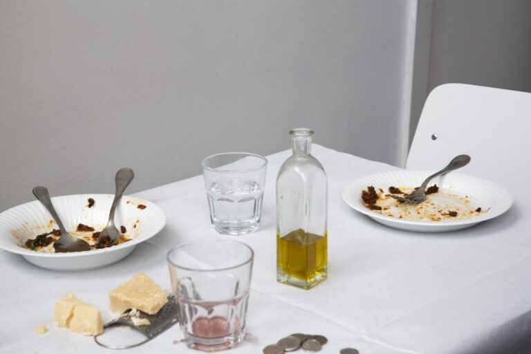 Auf einem Tisch mit weißem Laken sieht Man Teller mit Essensresten, ein angebrochenes Stück Peccorino und eine Karaffe Olivenöl – Symbolbild für Essensgebote und -verbote