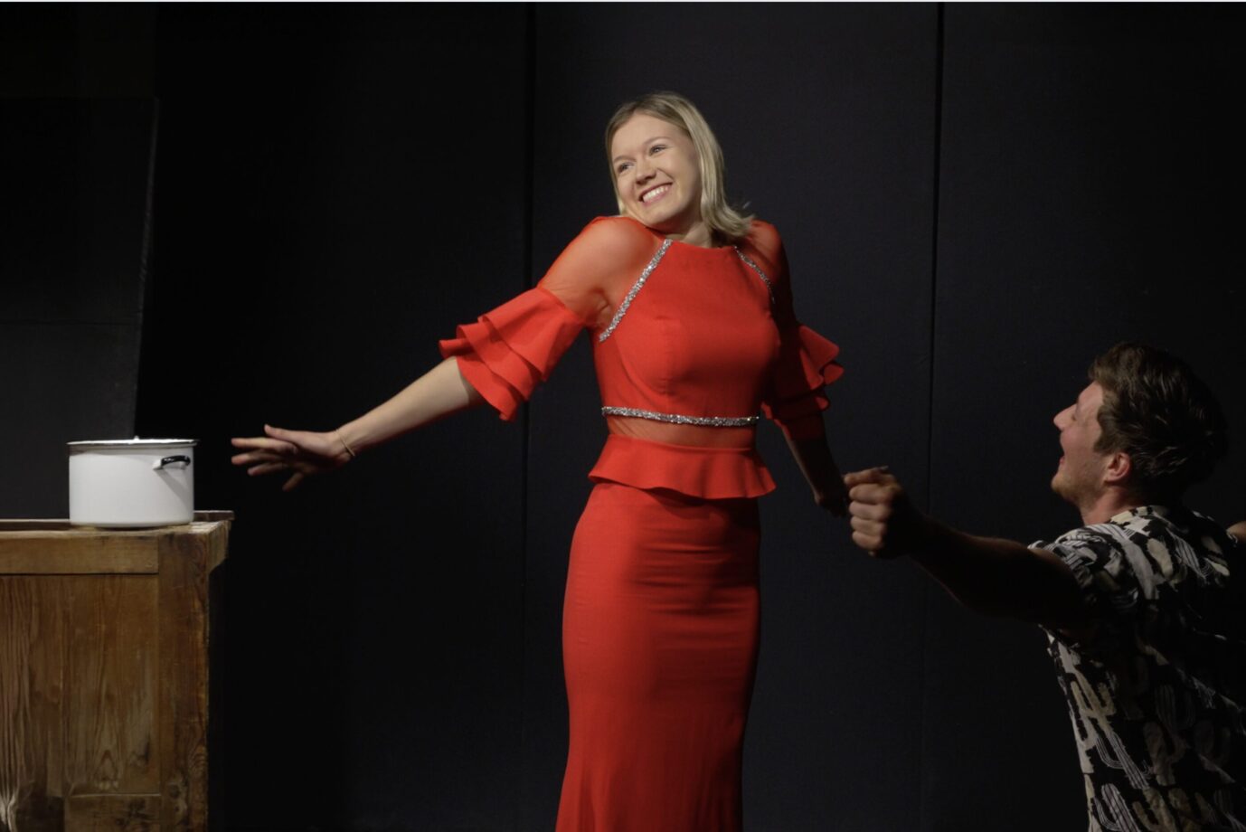 Vor einem schwarzen Hintergrund steht eine junge blonde Frau in einem roten Kleid und breitet ihre Arme aus – es handelt sich um die Autorin dieses Artikels, Michelle Gollan, auf einer Schulbühne in Berlin – Symbolbild für die ideologische Aufladung des deutschen Theaters