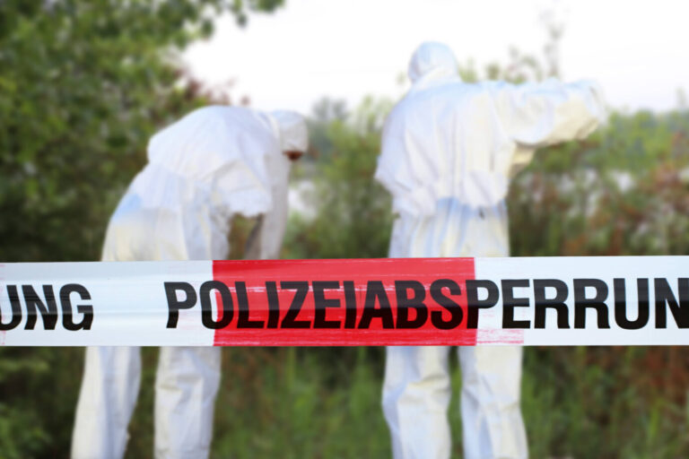 Im Vordergrund ist ein Absperrband der Polizei zu sehen, dahinter, leicht unscharf, sieht man zwei Polizisten in weißen Ganzkörperanzügen, die Spuren an einem Tatort sichern – Symbolbild für den zweifachen Mord, der nun in NRW von einem Ukrainer begangen wurde