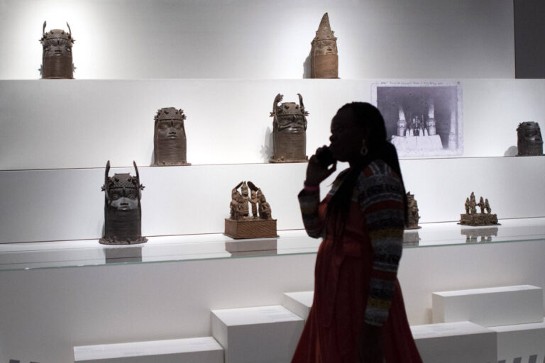 In einer weißen Vitrine stehen die Benin-Bronzen, davor steht eine schwarze Frau und betrachtet sie, Symbolbild für den Postkolonialismus