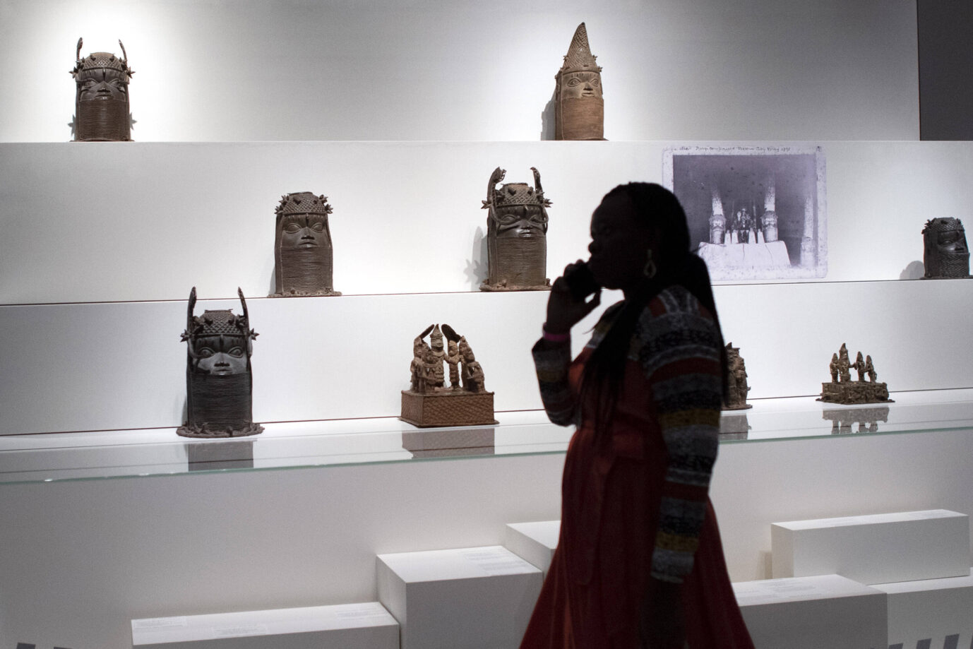 In einer weißen Vitrine stehen die Benin-Bronzen, davor steht eine schwarze Frau und betrachtet sie, Symbolbild für den Postkolonialismus
