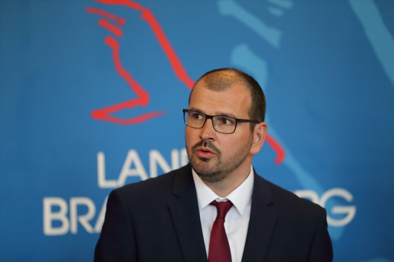 Der brandenburgische Bildungsminister Steffen Freiberg (SPD) steht vor einemk blauen Hintergrund auf dem "Land Brandenburg" steht – Freiberg verspricht, die Zahl der ausfallenden Unterrichtsstunden auf niedrigem Niveau zu halten und den Stundenausfall nicht eskalieren zu lassen.