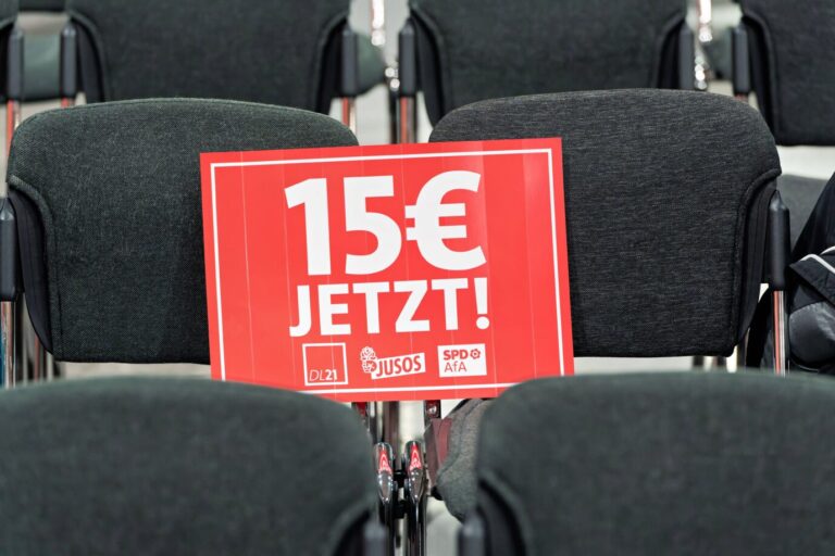 Ein rotes Schild mit der weißen Aufschrift „15 € JETZT!“ liegt auf einem gepolsterten, leeren Konferenzstuhl. Das Plakat zeigt die Logos von DL21, Jusos und SPD-Arbeitsgemeinschaft für Arbeitnehmerfragen (AfA). Es symbolisiert die Forderung der SPD-Jugendorganisation nach einem höheren gesetzlichen Mindestlohn. SPD-Wunsch knapp verfehlt: Der Mindestlohn steigt auf 14,60 Euro. Foto: IMAGO / Stefan Zeitz