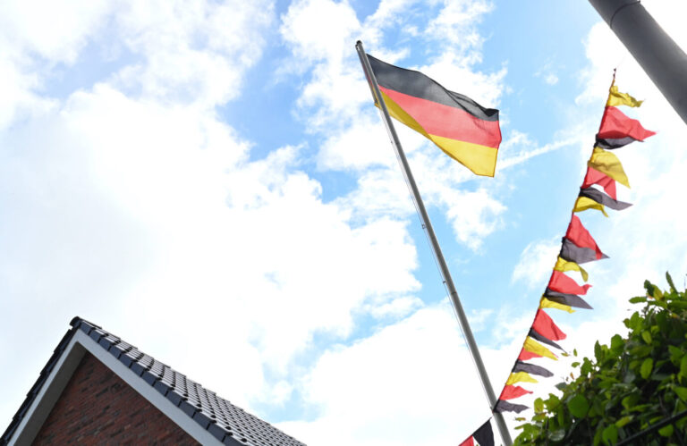Eine große Deutschlandflagge weht an einem Fahnenmast vor blauem Himmel mit vereinzelten Wolken. Rechts im Bild verläuft eine Wimpelkette mit kleinen Deutschlandfahnen. Links unten ist das spitze Dach eines roten Ziegelhauses mit dunkler Dachbedeckung zu sehen, am unteren rechten Bildrand ragen grüne Blätter ins Bild. Das Motiv zeigt eine typische Szene patriotischer Beflaggung im öffentlichen oder privaten Raum in Deutschland. Schwarz-Rot-Gold in Niedersachsen: Die CDU im Kreis Wolfenbüttel will die Deutschlandflagge dauerhaft hissen. Foto: IMAGO / diebildwerft