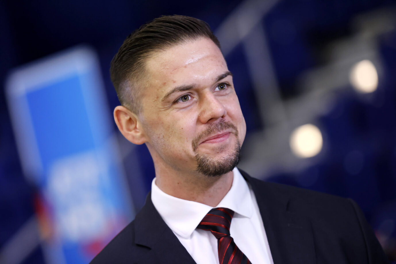 Hannes Gnauck, Bundestagsabgeordneter der AfD und Vorsitzender der Jungen Alternative, im Anzug mit weißem Hemd und rot-schwarzer Krawatte bei einer Veranstaltung. Er blickt leicht lächelnd zur Seite, im Hintergrund unscharf ein blauer Aufsteller mit AfD-Logo. Der AfD-Bundestagsabgeordnete Hannes Gnauck gut gelaunt: Disziplinarverfahren eingestellt.