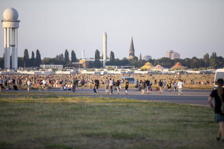 Man sieht eine grüne Wiese, darauf Menschen, dahinter Asphalt und im Hintergrund die Skyline Berlins – es handelt sich um das Tempelhofer Feld in Berlin – bald sollen hier Wohncontainer für Asylbewerber stehen.
