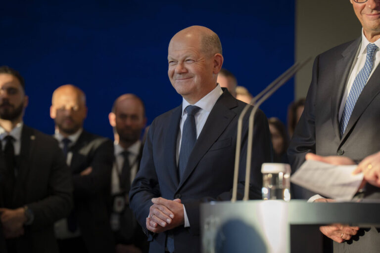 Bundeskanzler Olaf Scholz steht in einem dunklen Anzug lächelnd auf einer Bühne, im Hintergrund mehrere Männer in Anzügen. Die Szene wirkt offiziell, im Vordergrund ist ein Rednerpult mit Mikrofonen und einem Glas Wasser zu sehen. Scholz wird von Scheinwerfern beleuchtet, der Hintergrund ist in kräftigem Blau gehalten. Die Aufnahme vermittelt den Eindruck einer Pressekonferenz oder eines politischen Auftritts. Es ist die Amtsübergabe an Friedrich Merz. Der Christdemokrat führt nun eine schwarz-rote Bundesregierung an.