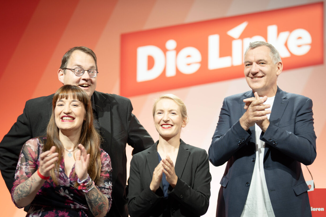 Vier Mitglieder der Parteispitze von Die Linke stehen nebeneinander auf der Bühne des Bundesparteitags 2025 in der Messe Chemnitz. Im Hintergrund ist groß das Logo der Partei zu sehen. Die Politiker – darunter Heidi Reichinnek, Sören Pellmann, Ines Schwerdtner und Jan van Aken – lächeln und klatschen. Alle wirken gelöst und zufrieden. Die Szene spielt sich in einem hell ausgeleuchteten Saal in Chemnitz, Sachsen, ab. Die Linkspartei erlebt ein Umfragehoch in Berlin. Bürgermeister Wegner besitzt keine Mehrheit und bleibt unbeliebt. Nur zwei Koalitionen sind rechnerisch möglich.