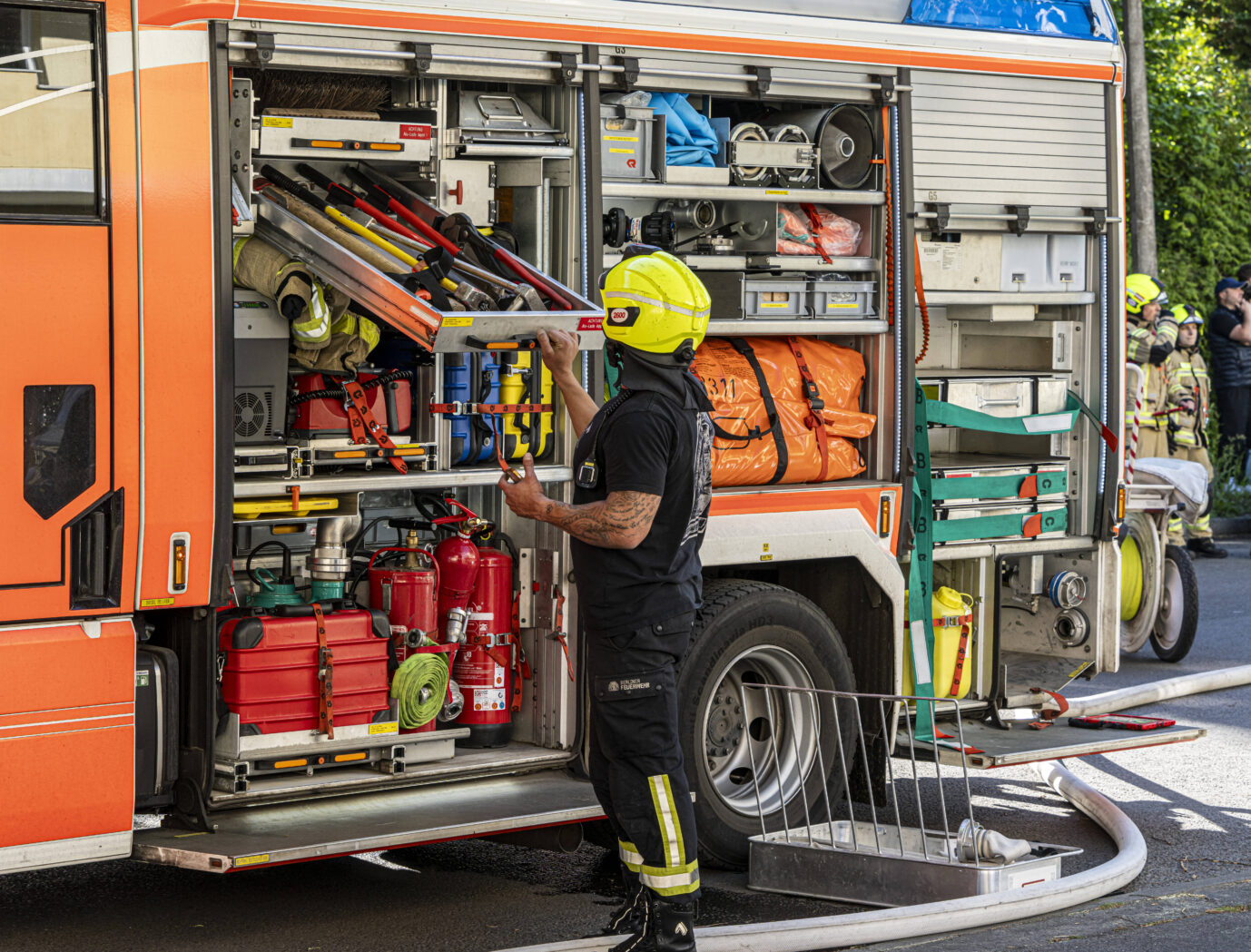 Ein Feuerwehrmann der Berliner Feuerwehr steht an einem geöffneten Einsatzfahrzeug und sortiert Ausrüstung. In der Seitenklappe des orange-weißen Löschfahrzeugs sind zahlreiche Geräte wie Äxte, Schläuche, Feuerlöscher und Notfalltaschen sichtbar. Der Feuerwehrmann trägt einen gelben Helm und eine schwarze Uniform mit reflektierenden Streifen. Im Hintergrund stehen weitere Einsatzkräfte in voller Schutzkleidung, einige beobachten das Geschehen. Die Szene spielt im öffentlichen Straßenraum bei Tageslicht in Berlin.