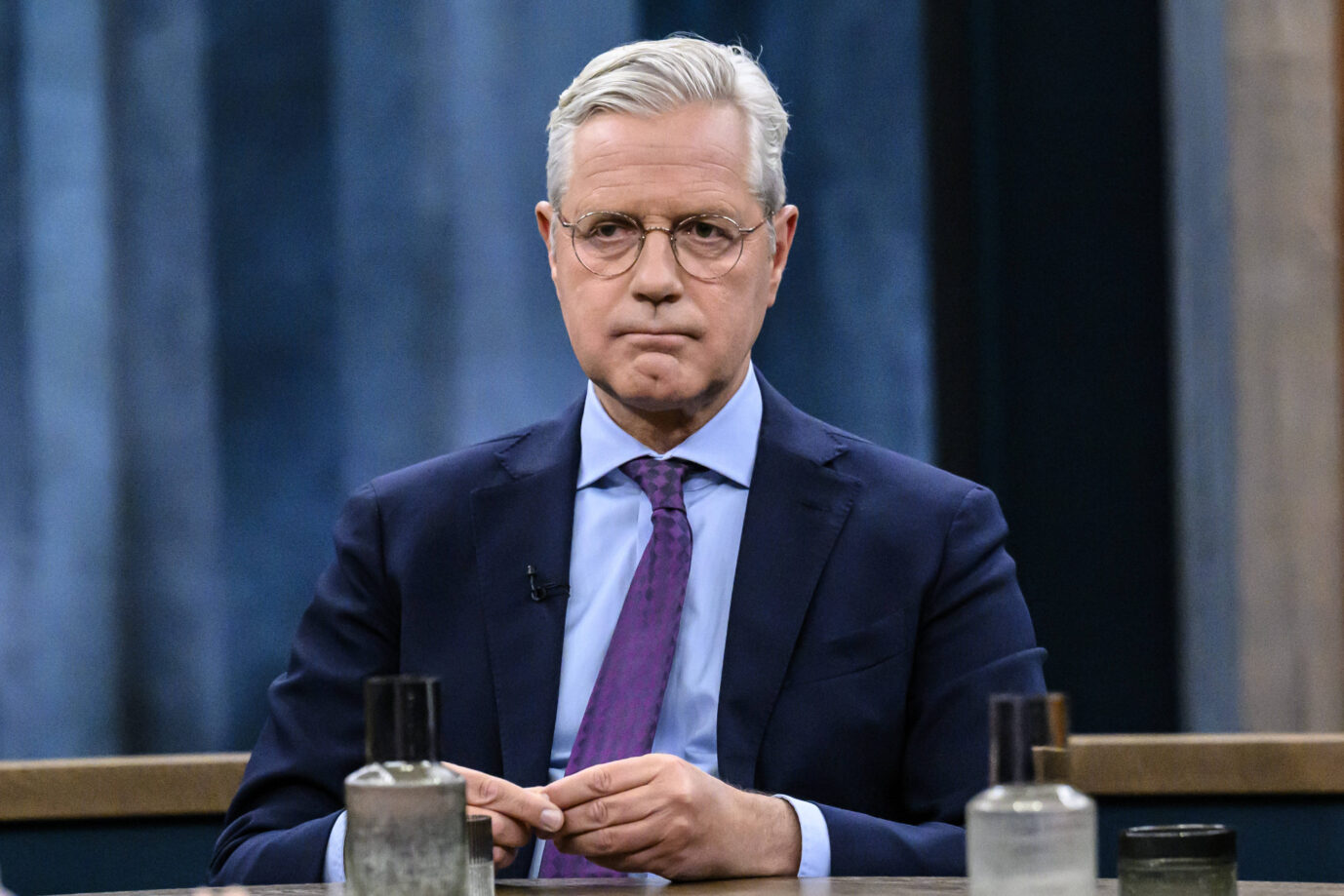 Norbert Röttgen, CDU-Außenpolitiker, mit ernster Miene in einem Fernsehstudio. Er trägt einen dunkelblauen Anzug, ein hellblaues Hemd und eine lilafarbene Krawatte. Im Vordergrund stehen mehrere Wassergläser und Flaschen auf einem Studiotisch. Die Bildszene wirkt konzentriert und sachlich. Das Bild eignet sich zur Bebilderung politischer Interviews oder außenpolitischer Debatten.