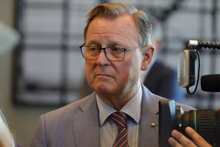 Bodo Ramelow, Bundestagsvizepräsident, mit ernstem Blick bei einer öffentlichen Veranstaltung im Berliner Reichstag. Er trägt ein hellblaues Sakko, ein Hemd und eine rot-blau gestreifte Krawatte; am Revers steckt ein Anstecker mit Deutschlandflagge. Ramelow steht vor unscharfem Hintergrund, eine professionelle Kamera mit Mikrofon und Licht ist im Vordergrund zu sehen. Anlass ist die Übergabe der Petition „phoenix muss bleiben!“, entgegengenommen von Bundestagspräsidentin Julia Klöckner.