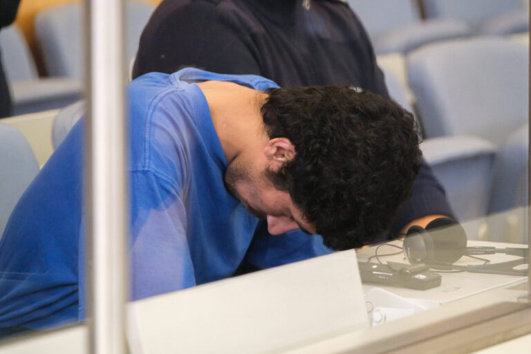Ein junger Mann mit dunklen Locken, in blauem T-Shirt, sitzt mit gesenktem Kopf in einem Gerichtssaal hinter einer Glasscheibe. Es handelt sich um den 27-jährigen Syrer und Asylbewerber Issa Al H., der am 27. Mai 2025 vor dem Düsseldorfer Oberlandesgericht erscheint. Ihm wird vorgeworfen, am 23. August 2024 auf einem Stadtfest in Solingen drei Menschen getötet und zehn weitere verletzt zu haben. Die Bundesanwaltschaft sieht eine islamistisch motivierte Tat und eine Mitgliedschaft in der Terrororganisation Islamischer Staat (IS). Auf dem Tisch vor ihm liegen Kopfhörer und Übersetzungstechnik. Das Bild dokumentiert den Prozess gegen den mutmaßlichen Attentäter.