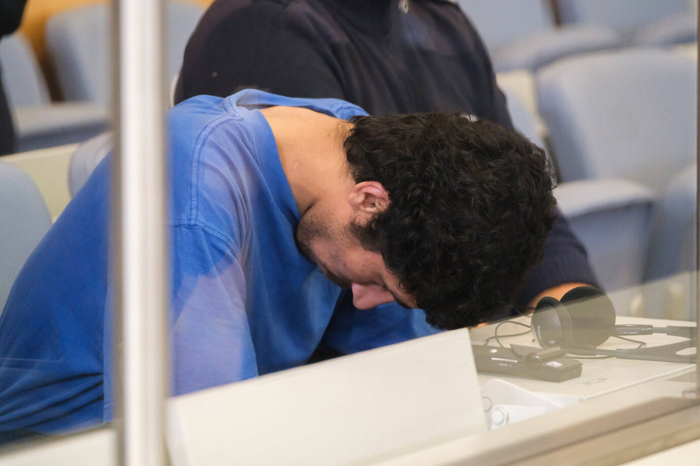Ein junger Mann mit dunklen Locken, in blauem T-Shirt, sitzt mit gesenktem Kopf in einem Gerichtssaal hinter einer Glasscheibe. Es handelt sich um den 27-jährigen Syrer und Asylbewerber Issa Al H., der am 27. Mai 2025 vor dem Düsseldorfer Oberlandesgericht erscheint. Ihm wird vorgeworfen, am 23. August 2024 auf einem Stadtfest in Solingen drei Menschen getötet und zehn weitere verletzt zu haben. Die Bundesanwaltschaft sieht eine islamistisch motivierte Tat und eine Mitgliedschaft in der Terrororganisation Islamischer Staat (IS). Auf dem Tisch vor ihm liegen Kopfhörer und Übersetzungstechnik. Das Bild dokumentiert den Prozess gegen den mutmaßlichen Attentäter.