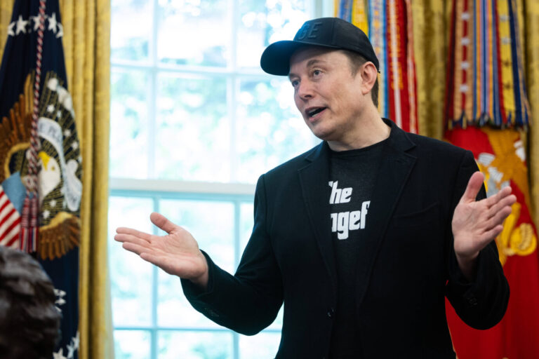 Elon Musk im Oval Office mit schwarzer Kappe, schwarzem Sakko und T-Shirt mit „Dogefather“-Aufdruck, gestikulierend vor US-Flaggen. Elon Musk bei seinem offiziellen Abschied im Weißen Haus – mit Doge-Cap und blauem Auge. Foto: IMAGO / ZUMA Press Wire