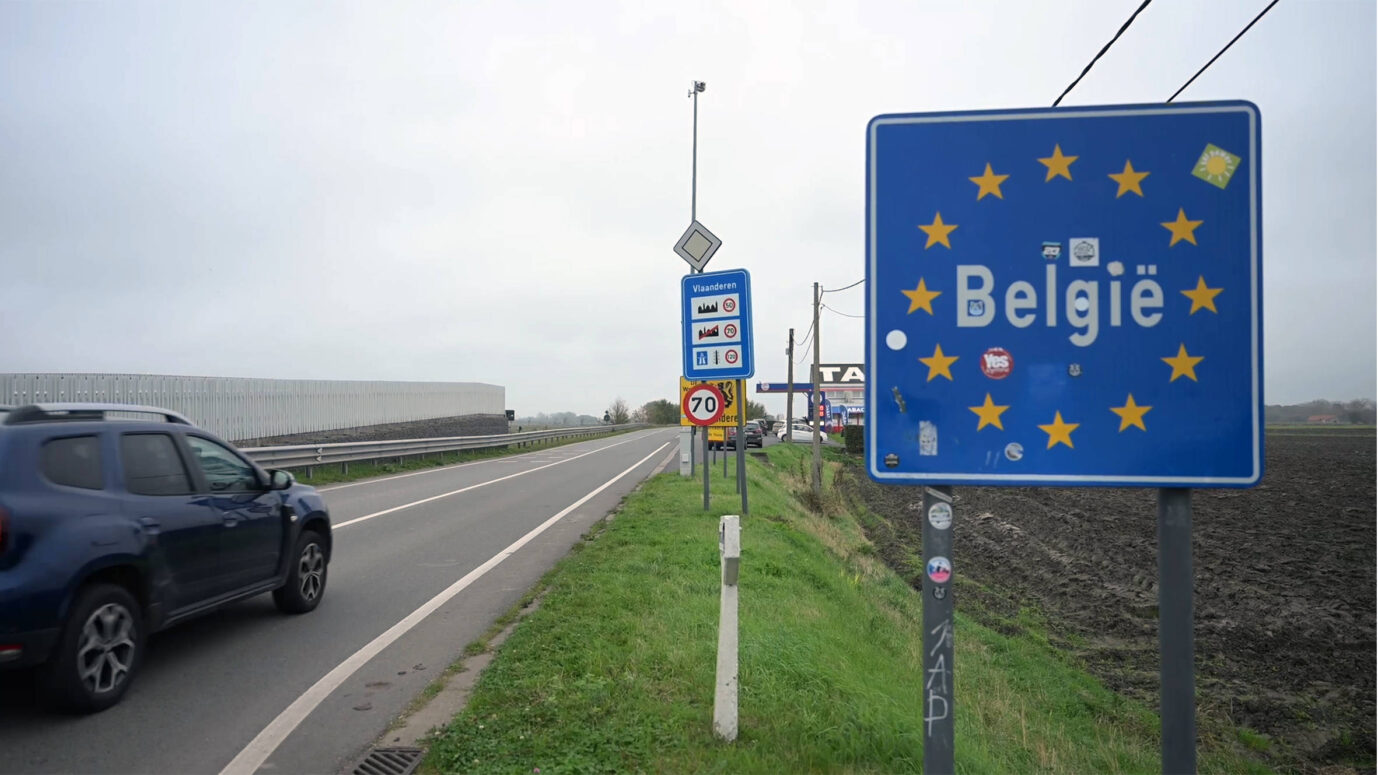 Straßenszene an der belgischen Grenze mit einem blauen Schild „België“ und zwölf gelben Sternen der EU, daneben weitere Verkehrsschilder, darunter ein Hinweis auf Flandern und ein Tempolimit. Ein Auto fährt in Richtung Landesinneres. Die Aufnahme entstand an einem bewölkten Tag und zeigt das flache Grenzgebiet mit Straße, Acker und Tankstelle. Grenzübertritt bei Flandern: Belgien will künftig wieder kontrollieren, wer ins Land fährt. Foto: IMAGO / Anadolu Agency