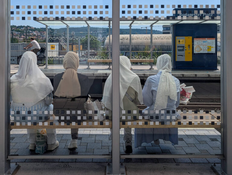 Vier junge Frauen mit hellen Kopftüchern sitzen auf einer Bank an einer überdachten Stadtbahnhaltestelle am Budapester Platz in Stuttgart. Sie blicken auf die gegenüberliegende Gleisseite, wo ein Fahrkartenautomat und ein Fahrplan hängen. Eine Frau hält einen Blumenstrauß mit roten und weißen Rosen. Im Hintergrund fährt ein Radfahrer vorbei, dahinter sind Hügel mit Wohnhäusern zu sehen. Das Bild zeigt eine Szene des Alltags im öffentlichen Nahverkehr. Vier Frauen an einer Haltestelle: Warten auf den Familiennachzug? Foto: IMAGO / NurPhoto