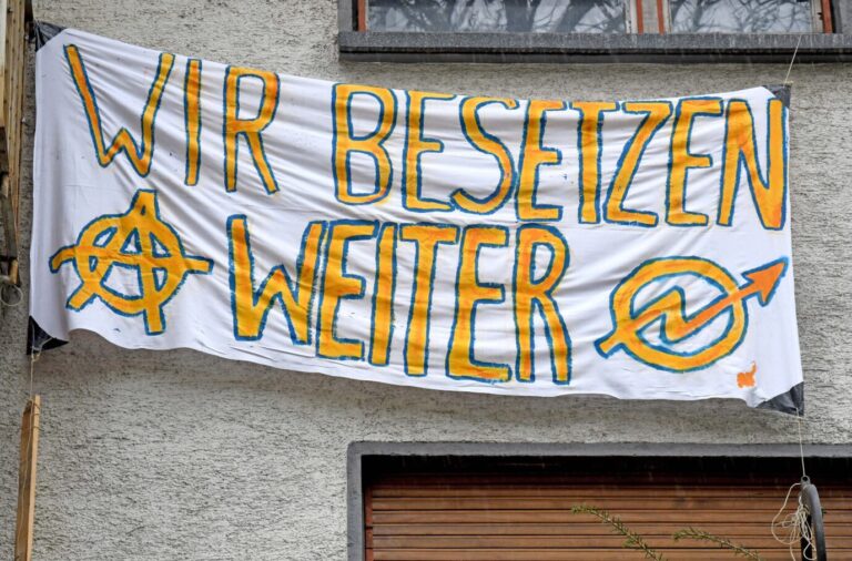 Auf einem weißen Transparent steht in gelber Schrift "Wir besetzen weiter", daneben ist das Logo der Hausbesetzerszene – Symbolbild dafür, daß sich im besetzten Haus RM16 in Dresden Linke gerade streiten