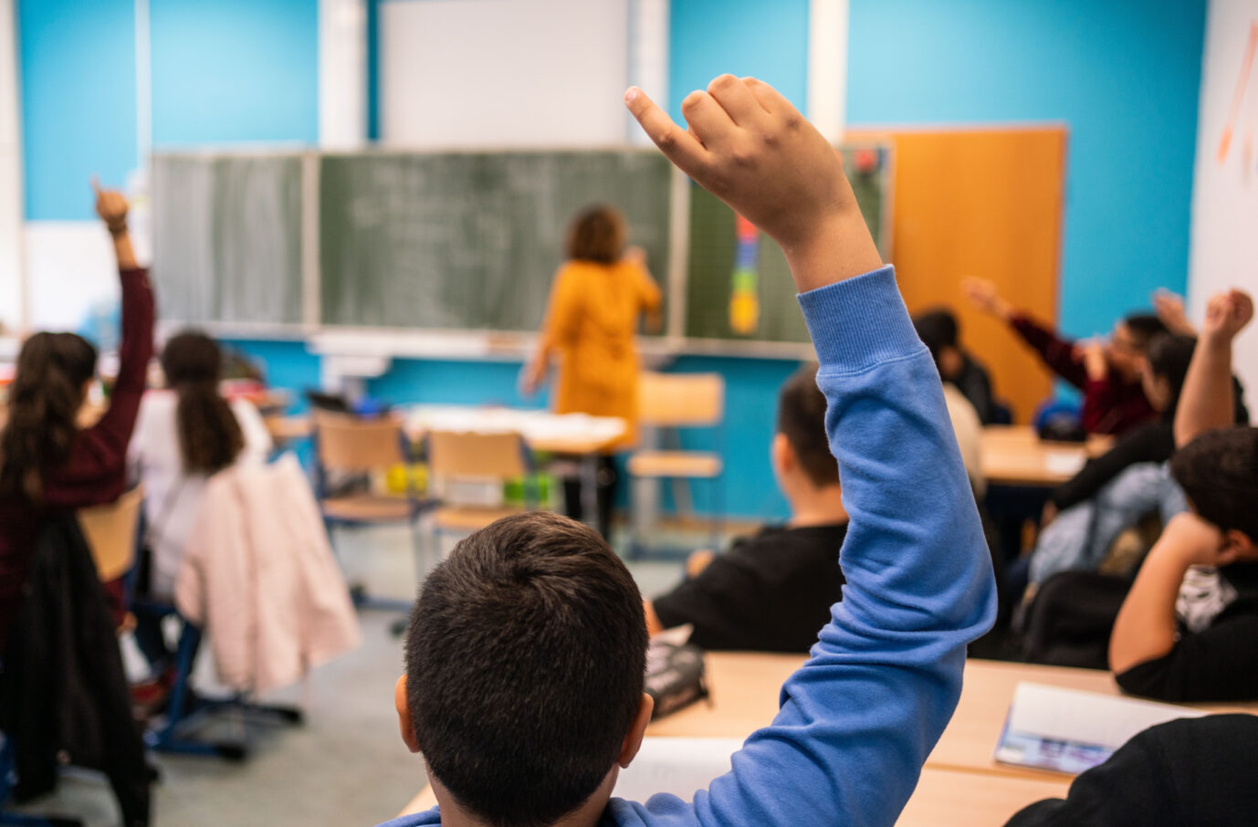 Schulklasse mit erhobenen Händen: Mehrere Schüler sitzen in einem bunt eingerichteten Klassenraum und melden sich. Im Vordergrund ist der Rücken eines Jungen mit blauer Kleidung zu sehen. An der Tafel steht eine Lehrerin in einem senfgelben Kleid und schreibt. Schüler melden sich im Unterricht: Blick in einen modernen Klassenraum mit Lehrerin an der Tafel und mehreren Kindern, die aktiv teilnehmen – Symbolbild für schulische Bildung und Unterrichtssituation. Islamunterricht an einer Frankfurter siebten Klasse: Verbände fordern ein flächendeckendes Angebot. Foto: picture alliance/dpa | Frank Rumpenhorst