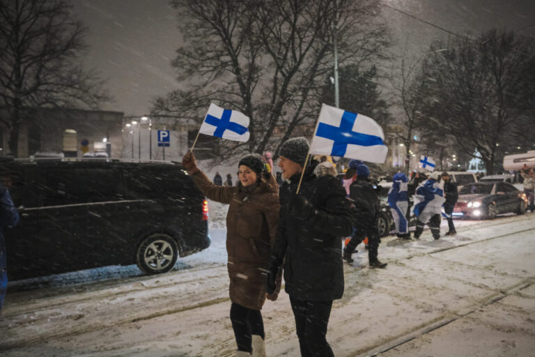 Auf einer verschneiten Straße in Helsinki halten zwei Finnen die finnische Fahne hoch – vielleicht sind sie rechts so wie mittlerweile die Mehrheit der Finnen