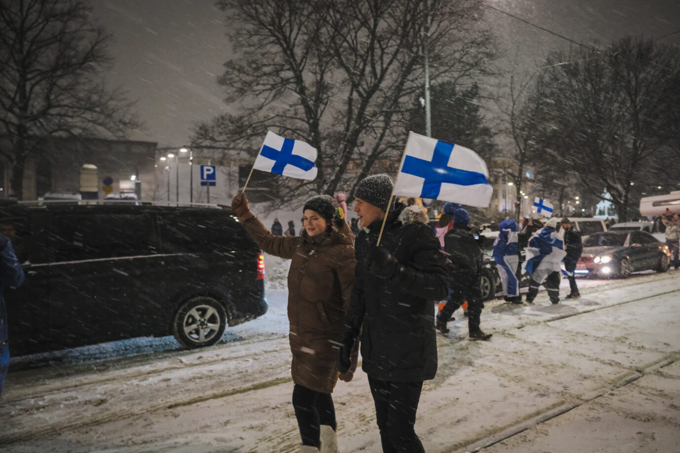 Auf einer verschneiten Straße in Helsinki halten zwei Finnen die finnische Fahne hoch – vielleicht sind sie rechts so wie mittlerweile die Mehrheit der Finnen