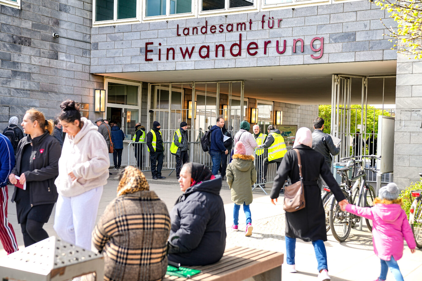 Ort der Einbürgerung: Menschen warten vor dem Landesamt für Einwanderung in Berlin. Sicherheitskräfte mit Warnwesten stehen vor dem Eingang, Besucherinnen mit Kopftuch und Kinderkleidung bewegen sich zum Gebäude. Im Vordergrund sitzen und stehen Personen in Winterkleidung. Schlange vor dem Landesamt für Einwanderung in Berlin: Beamte sollen gegen Bezahlung eingebürgert haben. Foto: picture alliance / SULUPRESS.DE | Vladimir Menck/SULUPRESS.DE